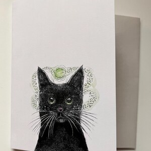 May include: Greeting card featuring a black cat with green eyes and long white whiskers. The cat has a detailed drawing style with a green and white halo-like design above its head. The card is white with a gray envelope.