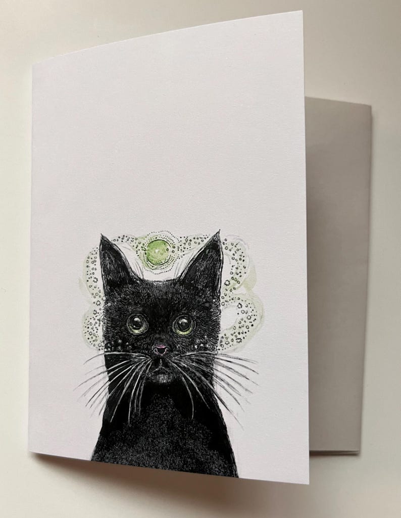 May include: Greeting card featuring a black cat with green eyes and long white whiskers. The cat has a detailed drawing style with a green and white halo-like design above its head. The card is white with a gray envelope.