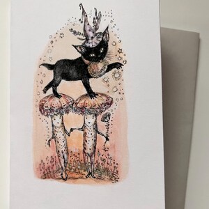 May include: Greeting card featuring a whimsical illustration. A black cat wearing a hat and collar stands atop two mushroom-headed figures. The card has a light pink and white background with a gray envelope.
