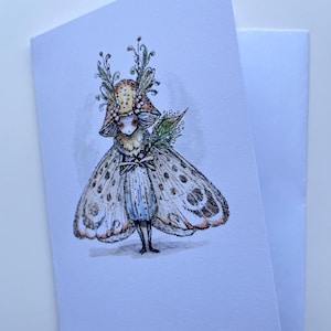May include: A folded greeting card featuring a whimsical illustration of a moth-like creature. The creature has large, patterned wings, a mushroom-shaped headpiece, and a blue dress. The artwork is detailed with soft colors and delicate lines, suggesting a fantasy theme.
