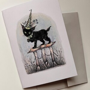 May include: Greeting card featuring a whimsical illustration of a black cat wearing a wizard hat, standing on three mushroom figures. The card has a light blue and gray background, with a matching envelope.