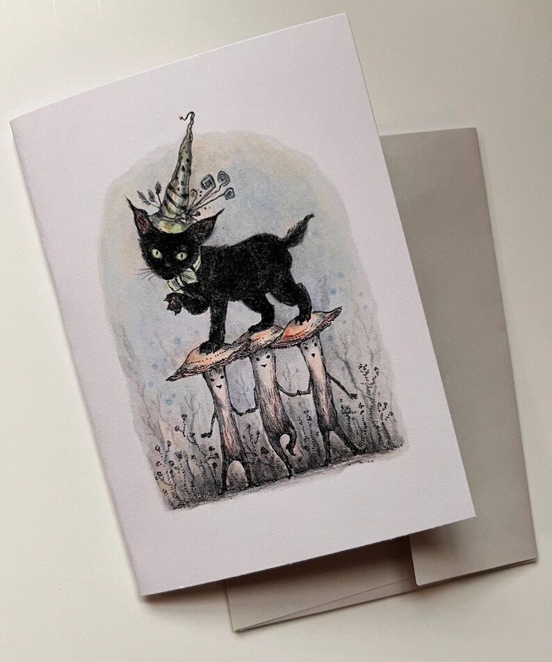 May include: Greeting card featuring a whimsical illustration of a black cat wearing a wizard hat, standing on three mushroom figures. The card has a light blue and gray background, with a matching envelope.