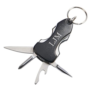 Op de afbeelding: Een zwarte multitool-sleutelhanger met een zilveren mes, een flesopener, een schroevendraaier en een klein puntig gereedschap. De sleutelhanger heeft een zilveren ring en de initialen "LJM" zijn gegraveerd op het handvat.