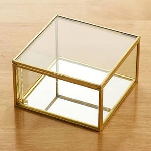 May include: A clear glass box with a gold-colored metal frame. The box has a hinged lid and a mirrored base, creating a reflective interior. The box is designed to hold small items.