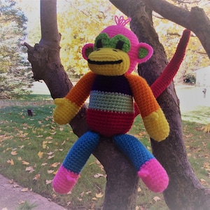 May include: A crocheted monkey toy with a rainbow striped body, pink head, green eyes, and a long tail. The monkey is sitting on a tree branch.