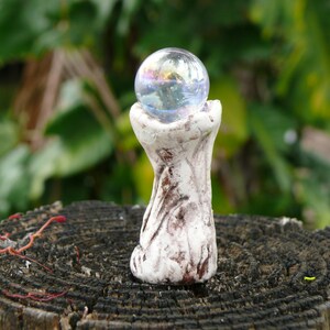 May include: A small, handcrafted sculpture featuring a clear, iridescent glass orb cradled by a white, textured ceramic base. The base has brown accents and sits atop a weathered wooden surface, with a blurred green foliage background.