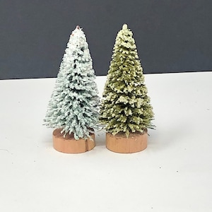 May include: Two miniature Christmas trees. One is covered in white flocking, the other is green with white flocking. Both trees are on small, round, wooden bases. The background is white and black.