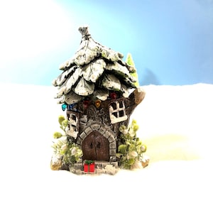 May include: A miniature Christmas-themed house with a snow-covered, leaf-shaped roof. The house has a stone facade, a wooden door, and small windows with colorful lights. A small red gift box sits on the steps. The house is surrounded by snow and greenery.