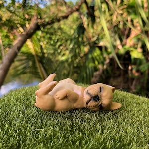 May include: A small, tan-colored figurine of a dog lying on its back with its eyes closed. The dog is positioned on a patch of green artificial grass, with a blurred green background of trees and foliage.
