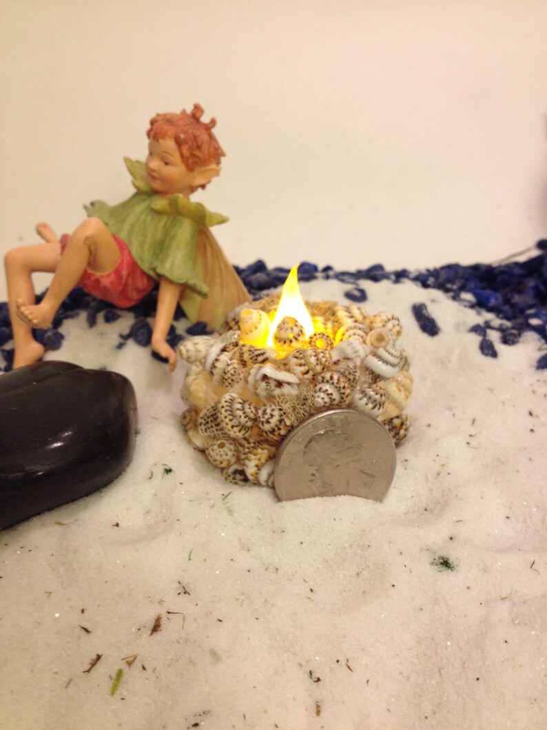May include: A small, round candle holder covered in tiny, speckled seashells with a glowing yellow LED flame. A silver US quarter coin is placed next to the holder for scale, alongside a small fairy figurine and a smooth black stone.