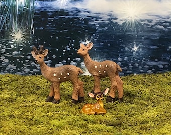 May include: Three miniature deer figurines. Two stand upright, with brown bodies and white spots. The third, smaller deer is lying down. The figurines are set on a bed of green moss, with a waterfall backdrop.