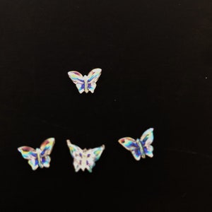 May include: Four iridescent butterfly-shaped embellishments. The butterflies have a shimmering, multi-colored effect, with hues of blue, green, and yellow. They are arranged against a dark background, highlighting their vibrant colors and intricate details.