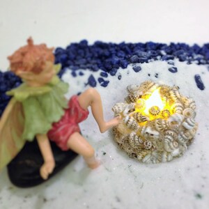 May include: A miniature fairy figurine with light brown hair, a green collar, and red shorts sitting on a black stone. Beside the figurine is a circular arrangement of small, speckled seashells surrounding a glowing, warm yellow LED light.