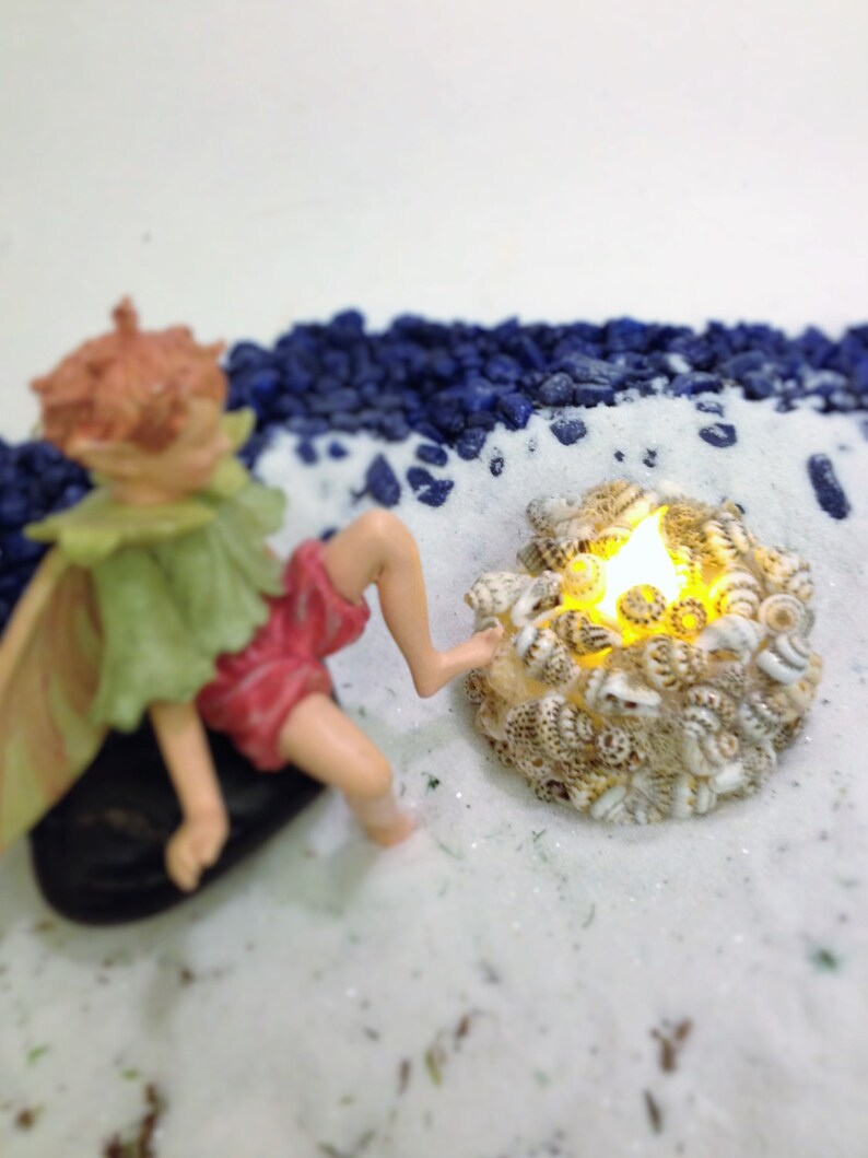 May include: A miniature fairy figurine with light brown hair, a green collar, and red shorts sitting on a black stone. Beside the figurine is a circular arrangement of small, speckled seashells surrounding a glowing, warm yellow LED light.