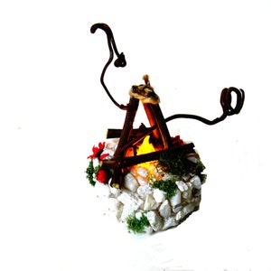 May include: A miniature campfire decoration featuring brown twigs arranged in a teepee shape over a glowing yellow light. The base is constructed from white stones, green moss, and small red accents, with two curved brown branches extending upward.