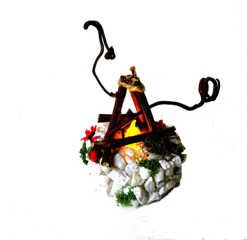 May include: A miniature campfire decoration featuring brown twigs arranged in a teepee shape over a glowing yellow light. The base is constructed from white stones, green moss, and small red accents, with two curved brown branches extending upward.