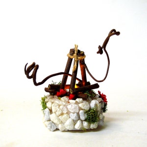 May include: A miniature campfire decoration featuring brown twigs arranged in a teepee shape, tied with twine, and set upon a circular base covered in white stones, green moss, and small red accents with a glowing yellow light.