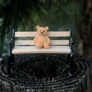 May include: A miniature tan teddy bear sits on a small wooden bench with black metal supports. The bench has light-colored wooden slats. The background is blurred, showing green foliage. The bear is centered on the bench, appearing to be resting.