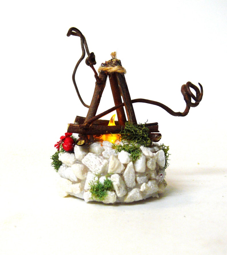 May include: A miniature campfire decoration featuring a circular base of white stones and green moss. A tripod structure made of brown twigs sits atop the stones, surrounding a small, glowing yellow LED flame light with red berries.
