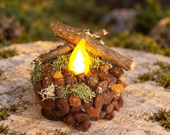 May include: A decorative campfire ornament constructed from small brown stones and green moss, featuring two crossed wooden twigs on top and a central, glowing yellow flame-shaped LED light that mimics the appearance of a small fire.