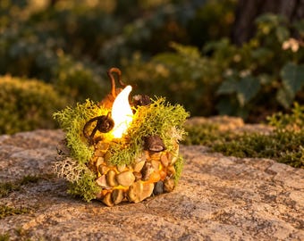 May include: A round decorative light fixture covered in small tan and gray pebbles, accented with bright green moss and dark brown curled twigs. A single warm white flame-shaped LED bulb glows from the center of the textured stone surface.