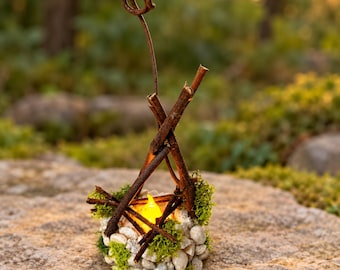 May include: A decorative campfire-style ornament constructed from small brown twigs arranged in a teepee shape, set upon a circular base of white pebbles and green moss, featuring a small, glowing yellow LED flame in the center.