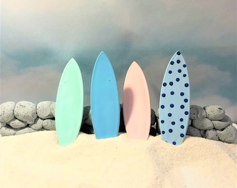 May include: Four colorful surfboards stand upright in the sand. The surfboards are pastel shades of green, blue, and pink, with one featuring a blue polka dot pattern. A stone wall and a cloudy sky form the backdrop.