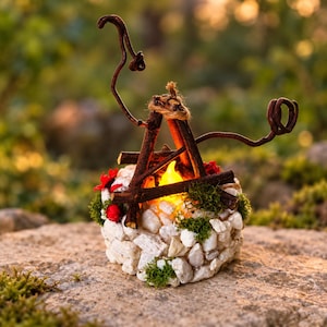 May include: A decorative miniature campfire ornament featuring a circular base of white stones and green moss. A small, glowing yellow LED flame sits inside a structure of dark brown twigs, accented with small red flowers and twine.