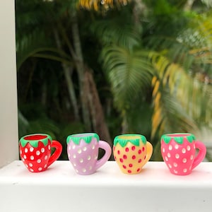 May include: Four small, colorful strawberry-themed mugs. The mugs are red, pink, yellow, and lavender, each with white polka dots and green accents. The mugs have small handles and are set against a blurred green background.