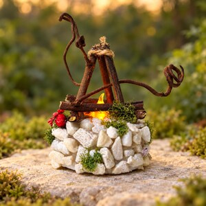 May include: A decorative miniature campfire ornament featuring white stones arranged in a circular base, topped with dark brown twigs arranged in a teepee shape, green moss accents, red berries, and a glowing yellow LED flame light.