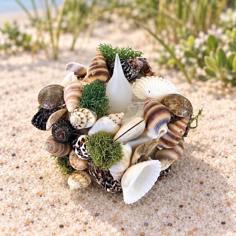 May include: A spherical decorative object covered in a variety of natural seashells in shades of brown, tan, and white, interspersed with patches of bright green preserved moss and featuring a central white teardrop-shaped flame-like accent.