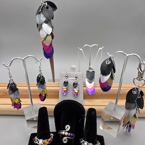 May include: A collection of handmade jewelry, including earrings, rings, a necklace, and a keychain. The pieces feature a scale-like design in various colors, including black, gray, silver, gold, pink, purple, and white. The jewelry is displayed on stands.