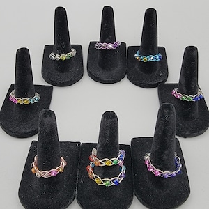 May include: A collection of colorful rings displayed on black velvet ring holders. The rings are made of intertwined wire and feature a variety of rainbow-colored beads. The text "Sexuality Rings" is at the top of the image.