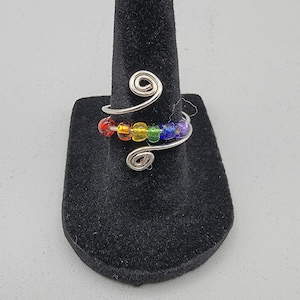 May include: A silver-toned wire ring with a spiral design, adorned with a row of colorful beads in rainbow hues. The word "Pride" is displayed below the ring.