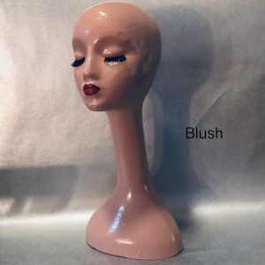 May include: A blush-colored mannequin head with closed eyes, long black eyelashes, and red lips. The head is on a long, slender neck and a rounded base. The word "Blush" is printed on the right side.