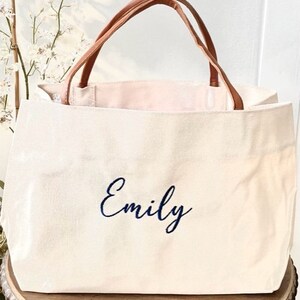 May include: Cream-colored tote bag with brown leather handles. The word "Emily" is embroidered in navy blue script on the front. The bag is sitting on a wooden table.