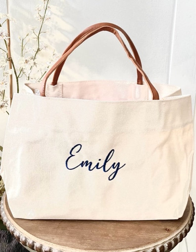 May include: Cream-colored tote bag with brown leather handles. The word "Emily" is embroidered in navy blue script on the front. The bag is sitting on a wooden table.