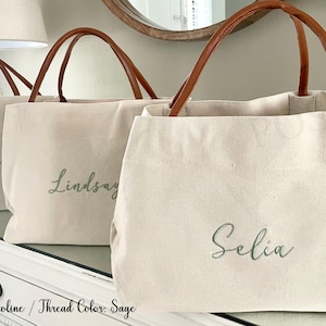 May include: Three canvas tote bags with brown leather handles. The bags are personalized with the names "Taylor", "Lindsay", and "Selia" in a sage green font. The font style is Caroline.