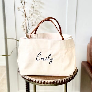 May include: A white canvas tote bag with brown leather handles. The bag has the name "Emily" embroidered on the front in black.