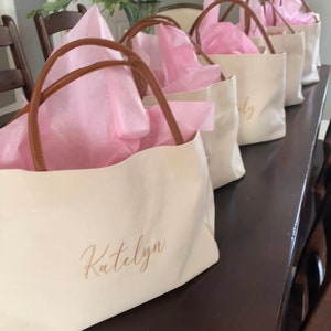 May include: Cream-colored tote bags with brown handles are arranged in a row. Each bag is filled with pink tissue paper. The name "Katelyn" is printed on the front of the first bag in a gold script.