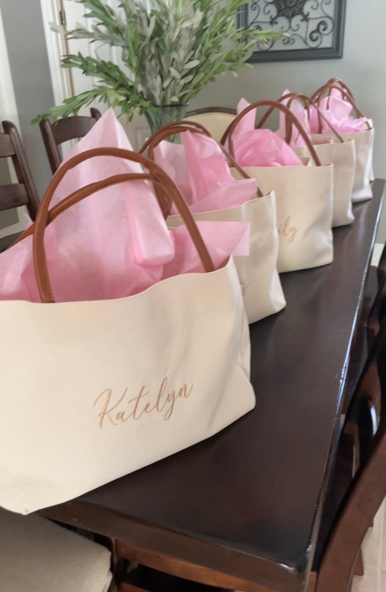 May include: Cream-colored tote bags with brown handles are arranged in a row. Each bag is filled with pink tissue paper. The name "Katelyn" is printed on the front of the first bag in a gold script.