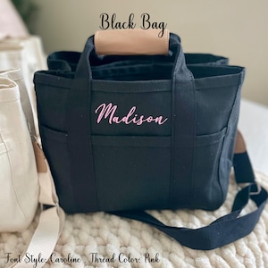 May include: Black tote bag with the name "Madison" embroidered in pink script. The bag has multiple pockets, a tan leather handle, and a black shoulder strap. The text "Black Bag" is above the bag.