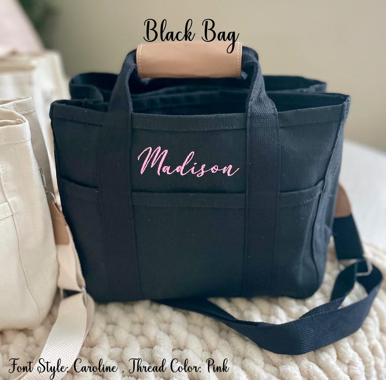 May include: Black tote bag with the name "Madison" embroidered in pink script. The bag has multiple pockets, a tan leather handle, and a black shoulder strap. The text "Black Bag" is above the bag.