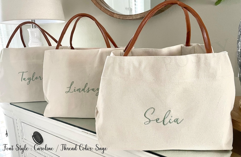 May include: Three canvas tote bags with brown leather handles. Each bag is a natural color and features a name embroidered in a sage green thread. The names are "Taylor", "Lindsay", and "Selia". The bags are sitting on a white dresser.