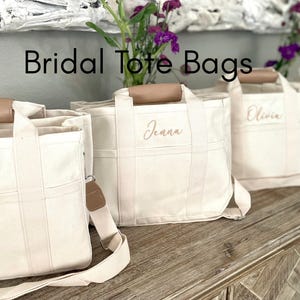 May include: Four off-white canvas tote bags with brown leather handles. Each bag has a different name embroidered on the front in a light pink color: Ella, Jenna, Olivia, and Emma. The bags are arranged on a wooden surface. Bridal Tote Bags.