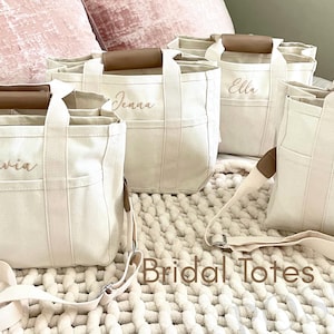 May include: Off-white canvas tote bags with brown leather handles and straps. Each bag is personalized with a name in a cursive font. The bags are labeled "Olivia", "Jenna", "Ella", and "Merria". The words "Bridal Totes" are printed on the bottom.