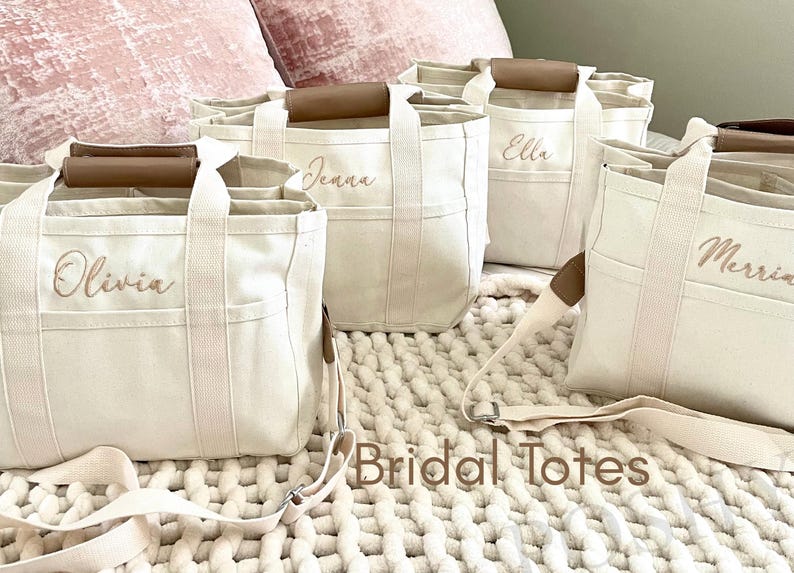 May include: Off-white canvas tote bags with brown leather handles and straps. Each bag is personalized with a name in a cursive font. The bags are labeled "Olivia", "Jenna", "Ella", and "Merria". The words "Bridal Totes" are printed on the bottom.
