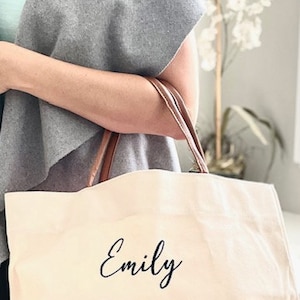 May include: A white canvas tote bag with brown leather handles. The bag has the name "Emily" printed in black cursive on the front.