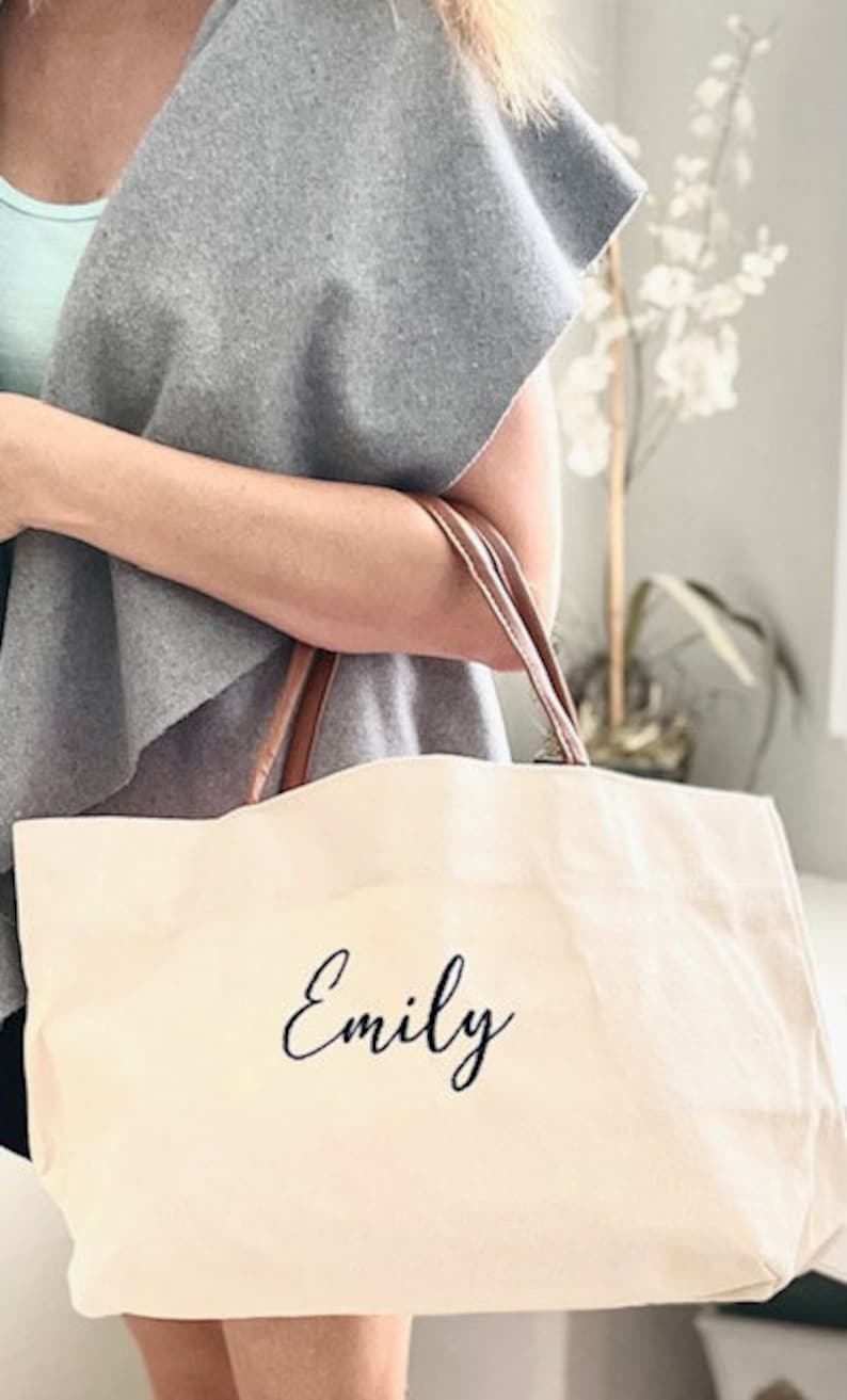 May include: A white canvas tote bag with brown leather handles. The bag has the name "Emily" printed in black cursive on the front.