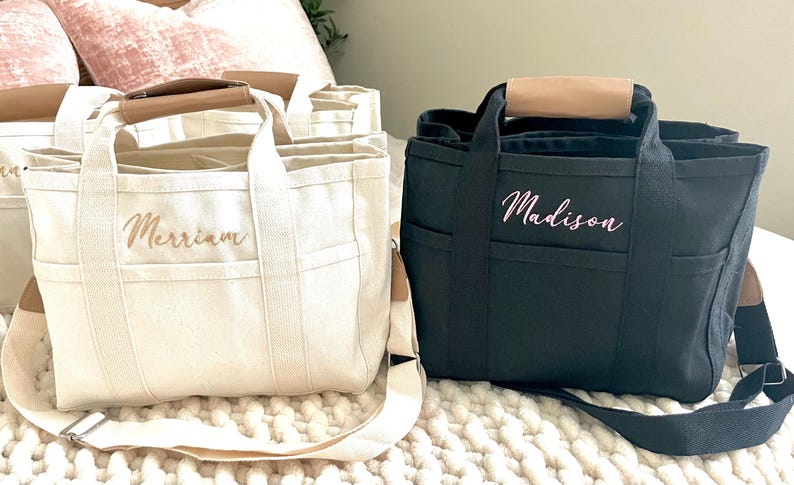 May include: Canvas tote bags in cream and black, featuring brown leather handles and shoulder straps. The cream bag has "Merriam" embroidered, while the black bag says "Madison". These bags have multiple pockets and are ideal for everyday use.